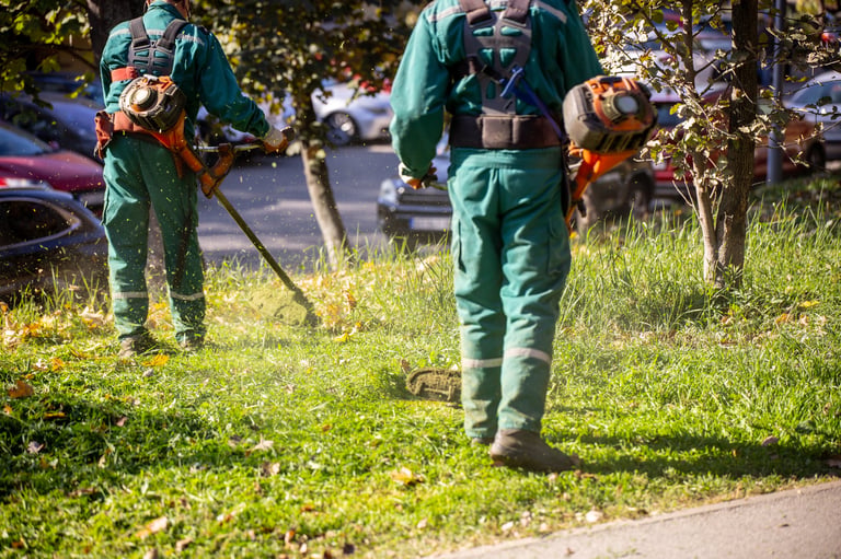 Two landscapers in green uniforms trimming grass in a city park